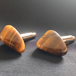 May include: A pair of cufflinks featuring polished tiger's eye stones. The stones are a warm brown color with golden striations, set on silver-toned metal posts. The cufflinks are displayed on a dark, textured surface.