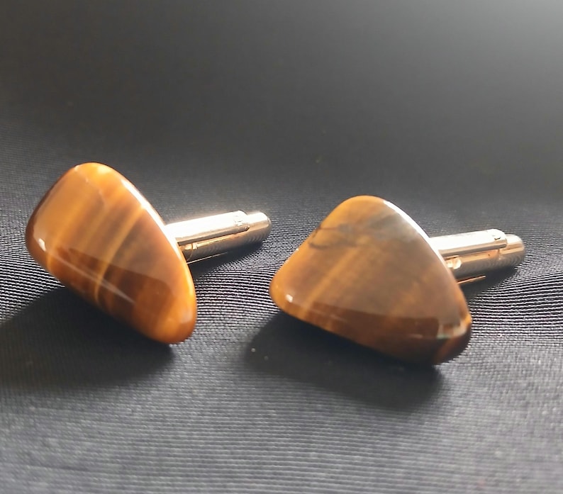 May include: A pair of cufflinks featuring polished tiger's eye stones. The stones are a warm brown color with golden striations, set on silver-toned metal posts. The cufflinks are displayed on a dark, textured surface.