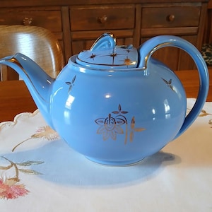 May include: A light blue ceramic teapot with gold trim and decorative gold floral and starburst designs. The teapot has a curved handle and a lid with a small handle. The teapot is on a white tablecloth.