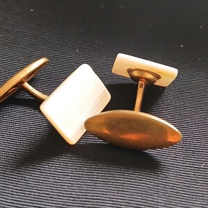 May include: A pair of vintage cufflinks with rectangular mother-of-pearl faces and gold-toned metal settings. One cufflink is positioned with the face up, while the other is turned to show the oval backing.