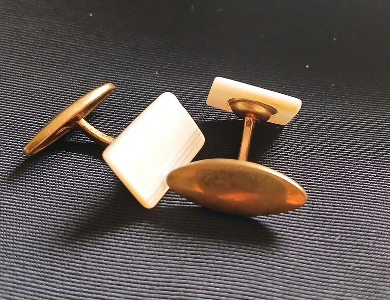 May include: A pair of vintage cufflinks with rectangular mother-of-pearl faces and gold-toned metal settings. One cufflink is positioned with the face up, while the other is turned to show the oval backing.