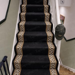 Op de afbeelding: Zwarte traploper met een luipaardprint border. De loper is geïnstalleerd op een houten trap met witte balustrades. De luipaardprint heeft bruine en beige vlekken op een crèmekleurige achtergrond. De tekst "Tapingrug" is zichtbaar.