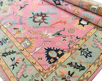 Könnte beinhalten: Eine aufgerollte rosa Teppich mit einem detaillierten Muster. Der Teppich hat ein zentrales rosa Feld mit verschiedenen Formen in Hellblau, Braun und Gelb. Ein Rand in Hellgrün und Gelb umrahmt das Design. Der Teppich hat einen Fransen.