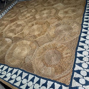 May include: A large, rectangular jute rug with a geometric pattern. The rug features a repeating pattern of interconnected circles and hexagons in shades of brown and tan. A border of blue and white geometric shapes frames the rug.