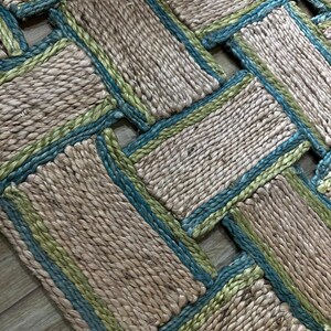 May include: Close-up of a woven rug with a natural fiber appearance. The rug features a rectangular pattern with a beige base, accented by teal and green borders. The texture appears coarse and handmade, suitable for home decor.