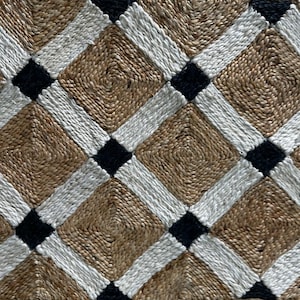 May include: A woven rug with a geometric pattern. The rug features a natural tan background with a repeating pattern of white and black diamond shapes. The rug is made of a natural fiber material.