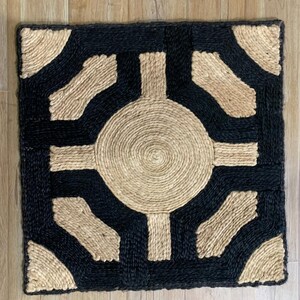May include: Square woven rug with a geometric design. The rug features a central circular pattern and corner accents in a natural tan color, framed by a black border and connecting shapes. The rug is made of a natural fiber material.
