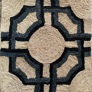May include: Square woven jute pillow with a geometric design. The design features a central circular element and intersecting lines in black and dark blue, creating a striking contrast against the natural jute color.