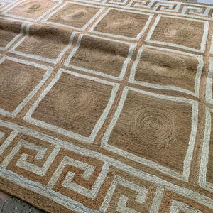 May include: A rectangular jute rug with a geometric design. The rug features a repeating pattern of squares and circles in shades of brown and beige. The border has a Greek key design.