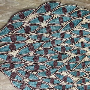 May include: A decorative, oval-shaped rug with a peacock feather design. The rug features teal, brown, and beige woven fibers. The design includes teal leaf shapes with brown circular accents, creating a textured surface.