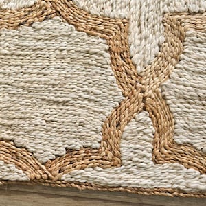 May include: Close-up of a natural fiber rug with a geometric pattern. The rug features a light beige central field with a repeating star-like design outlined in a darker tan color. The texture appears woven and slightly raised.