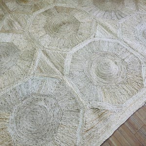 Puede incluir: Una alfombra tejida con un patrón geométrico repetitivo de grandes formas octogonales conectadas por triángulos más pequeños. La alfombra está construida con fibras naturales de color beige, mostrando un diseño texturizado y enrollado.