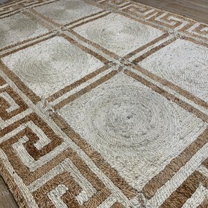 May include: A rectangular jute rug with a geometric design. The rug features a repeating pattern of squares and Greek key borders in natural and brown tones. The center of each square has a circular design.