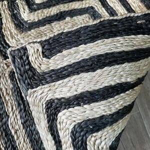 May include: Close-up of two woven jute rugs with a geometric pattern. The rugs feature alternating black and natural beige stripes, creating a bold, textured design. The edges are neatly finished, showcasing the craftsmanship of the rugs.