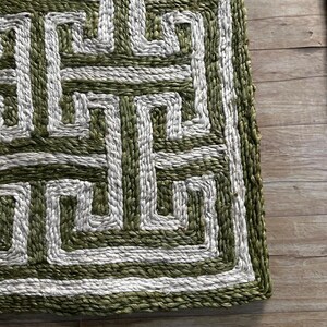 May include: Close-up of a rectangular rug with a geometric pattern. The rug features a repeating design in shades of green and white. The rug is made of woven material, with a textured appearance.
