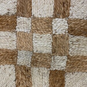 May include: Close-up of a woven rug with a checkerboard pattern. The rug features alternating squares of light beige and brown fibers, creating a textured surface. The natural tones suggest a rustic or bohemian style.