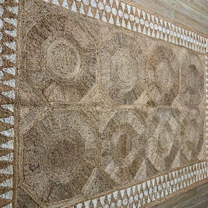 May include: A rectangular jute rug with a geometric design. The rug features large, circular patterns in the center, framed by a border of white triangles and circles. The rug's natural brown color and textured surface create a warm, inviting aesthetic.