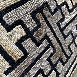 May include: Woven textile with a geometric pattern. The design features a repeating pattern of black, beige, and gray lines. The texture appears to be made of natural fibers, creating a rustic and handcrafted aesthetic. The overall shape is square.