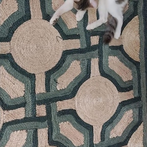 May include: A woven rug with a geometric pattern in shades of green and beige. The rug features circular and angular shapes. A cat with white and gray fur is lying on the rug.