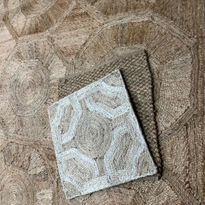 May include: A natural fiber rug with a geometric design. The rug features a large, circular pattern with a smaller, square rug on top. The colors are neutral, with shades of brown and beige. The texture appears to be woven.
