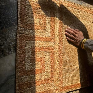 May include: Woven rectangular rug with a geometric design in shades of tan and orange. The rug is made of natural fibers and has a textured surface. The border is a darker shade of tan. The rug is approximately 36 inches long and 24 inches wide.