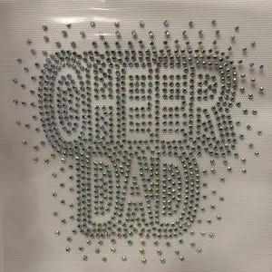 May include: A rhinestone design spelling out "CHEER DAD" on a white background. The rhinestones are iridescent, reflecting multiple colors. The design is likely for apparel or crafts.