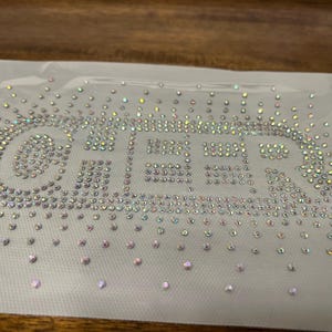 May include: A clear, rectangular sheet with the word "CHEER" spelled out in sparkling, iridescent rhinestones. The rhinestones are arranged to form the letters, with additional scattered rhinestones around the word.