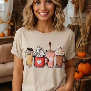 Puede incluir: Una mujer con una camiseta beige con una ilustración de cuatro bebidas de café. Las bebidas incluyen un batido con chispas de chocolate, un café con crema batida en una taza roja, una bebida rosa con pajita y un café con crema batida.