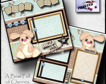 Può includere: Una pagina di scrapbooking con uno schema di colori blu e marrone. La pagina presenta un tema da bambino con una simpatica illustrazione di coniglio, una stella e il testo "Piccolo angelo". La pagina include anche il testo "Baby Boy" in lettere maiuscole e il testo "Una ciotola piena di ciliegie" in basso.