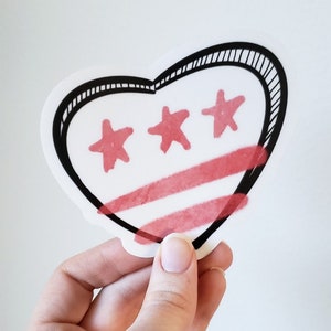 May include: A heart-shaped sticker with a black outline and a red and white design. The design features three red stars and three red stripes.