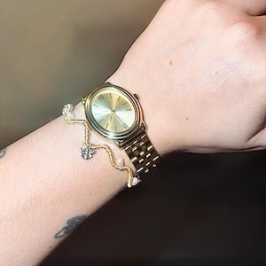 May include: Gold-toned wristwatch with a matching gold bracelet. The watch has a round face with Roman numeral hour markers and a linked band. The bracelet features a delicate design with small charms and pearls.