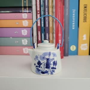 Può includere: Una piccola teiera in ceramica bianca con un paesaggio di mulino a vento dipinto in blu. La teiera presenta un manico in filo metallico azzurro e dettagli blu sul coperchio e sul beccuccio, creando un oggetto decorativo nei colori blu e bianco.