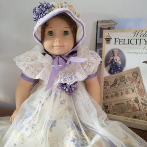 Pode incluir: Uma boneca de cabelos castanhos e olhos verdes usa uma touca branca com detalhes florais roxos e uma fita roxa. A boneca está vestida com um vestido branco com acabamento em renda e uma sobreposição transparente com um padrão floral roxo e verde.