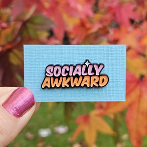 Puede incluir: Un pin con la frase "Socially Awkward" en letras rosas y naranjas con contorno negro, sobre un fondo azul claro. El fondo muestra hojas otoñales de colores.