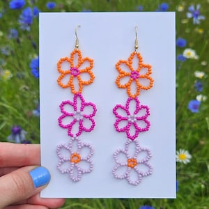 May include: A pair of dangle earrings with three beaded flower charms. The top flower is orange, the middle flower is pink, and the bottom flower is light purple.