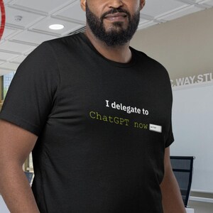 Puede incluir: Camiseta negra con el texto "I delegate to ChatGPT now." Un sello circular rojo dice "XL APPROVED FINEST CHAOS BUILT FOR MORE." La persona lleva vaqueros azules en un entorno de oficina.