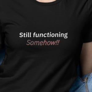 Puede incluir: Camiseta negra de cuello redondo con el texto "Still functioning Somehow!!" en blanco y rosa. Una prenda de vestir informal.