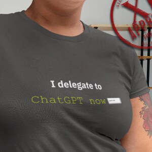 Puede incluir: Camiseta gris oscuro con el texto "I delegate to ChatGPT now" en blanco y verde. La camiseta es de talla XL y tiene un sello redondo rojo que dice "APPROVED". La persona lleva un cinturón negro con hebilla dorada.