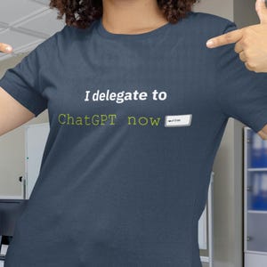 May include: Dark blue t-shirt with the text "I delegate to ChatGPT now" in white and green. The shirt is being pointed at by a person.
