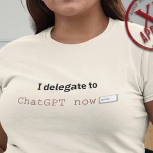 May include: Cream-colored t-shirt with the text "I delegate to ChatGPT now" in black and brown. The shirt has a crew neck and short sleeves. A red stamp that says "XL Approved" is in the upper right corner.