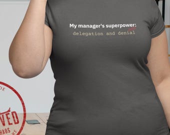 May include: Dark gray t-shirt with the text "My manager's superpower: delegation and denial" in white and pink. A red XL approved stamp is visible on the left side of the image.