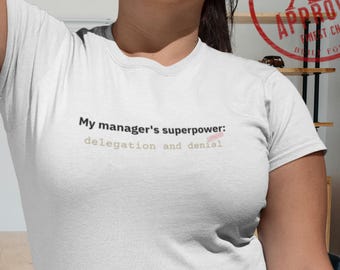 May include: White t-shirt with the text "My manager's superpower: delegation and denial" in a casual font. A red stamp with the words "XL APPROVED" is visible in the upper right corner. The shirt is worn with blue jeans.