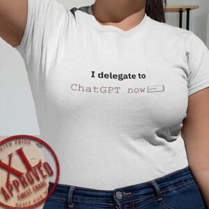 Puede incluir: Camiseta blanca con el texto "I delegate to ChatGPT now" en negro y gris. Un sello circular rojo y marrón dice "XL Approved, Finest Chaos, Built for M, Sized with Pride."