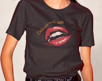 May include: A black t-shirt with a graphic of red lips and the gold text "Delicate with teeth". The lips are detailed with shading and highlights, and the teeth are white. The t-shirt is a crew neck style.