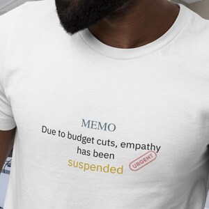 May include: White t-shirt with the text "MEMO Due to budget cuts, empathy has been suspended" in a mix of colors. A red "URGENT" stamp is also visible. The shirt is a crew neck style.