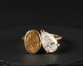 May include: A gold ring featuring two gemstones: an oval-cut, light-yellow stone and a pear-shaped, clear stone. The ring is set on a dark, round surface against a black background. The ring's design is elegant and modern.