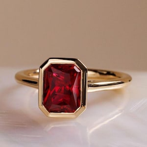 Op de afbeelding: Een gouden ring met een rechthoekige, dieprode edelsteen. De edelsteen is gezet in een gouden bezel en de band is van gepolijst goud. De ring wordt getoond op een wit oppervlak, met een zachte, neutrale achtergrond.