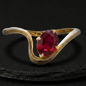 Puede incluir: Un anillo de oro con un rubí rojo intenso de talla ovalada en un engaste de garras. La banda tiene un diseño liso, curvo y ondulado que fluye alrededor de la piedra central, creando una forma fluida y continua en oro amarillo pulido.