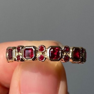 May include: A gold ring with a series of bright red gemstones. The ring's design includes both square and round-cut rubies, creating a striking contrast. The gemstones are set in a gold band, showcasing a classic and elegant style.