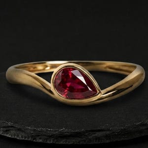 Puede incluir: Un anillo de oro con una sola piedra preciosa roja tallada en forma de pera y facetada, engastada en una banda lisa y pulida. La banda se curva suavemente alrededor de la base de la piedra, creando una silueta fluida y continua.